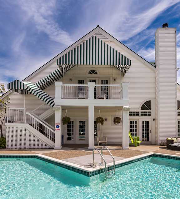 A white house with a striped awning and a pool in front.