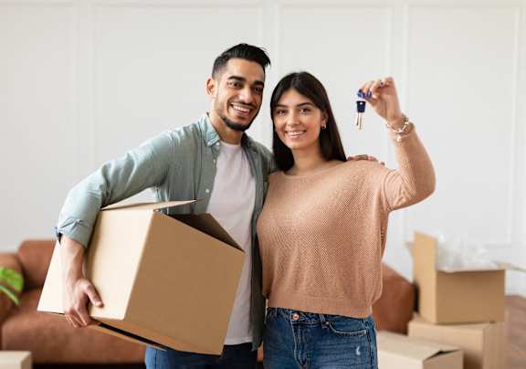 A man and woman are standing in a room with boxes, holding a key and a box.