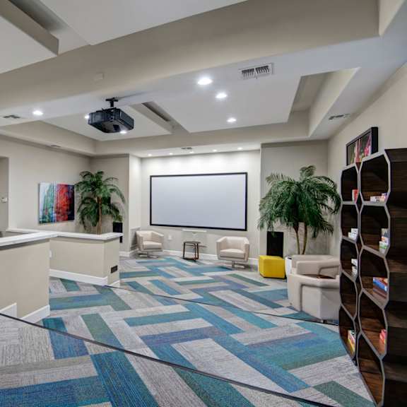 The common room at Rio Paradiso with plush chairs, a TV, plants, and bookshelves with board games on them at Rio Paradiso Apartment Homes. 