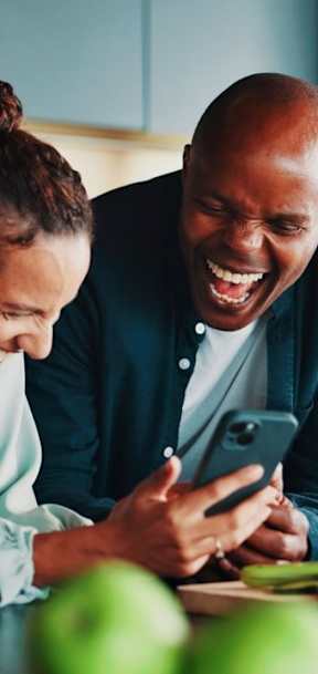 A man and woman are laughing while looking at a phone in a kitchen.