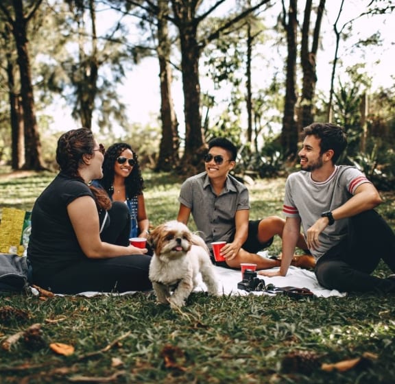 A group of people are sitting on a blanket in a park with a dog. at River Walk Apartments, Idaho