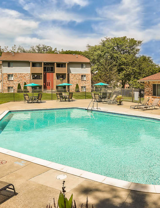 Outdoor swimming pool with expansive sundeck with lounge chairs. Exterior of Brighton Cove can be seen in the background. A pool house is off to the right. 