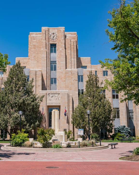 A large, beige-colored building with a clock on the front is surrounded by trees and has a walkway in front.
