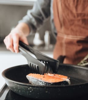 a man cooking salmon in a pan with a spatula at The Commons at Rivertown, Grandville, MI