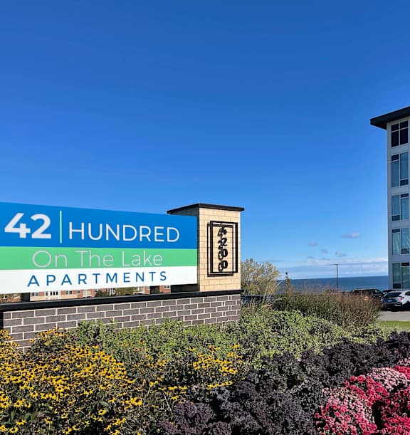 A modern apartment building is situated by the water with a clear blue sky above at 42 Hundred On The Lake, St Francis, WI