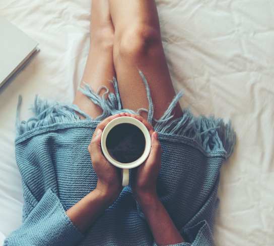 A person is lying on a bed with their legs up, holding a cup of coffee and a book.