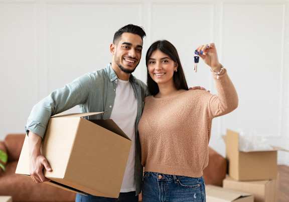 A man and woman are standing in a room with boxes, the man is holding a box and the woman is holding a key.