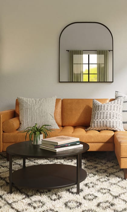 Brown leather couch with white and cream throw pillows. In front of the couch is a round coffee table with books and a plant resting on it. A white rug with a balck desgin is under the couch.