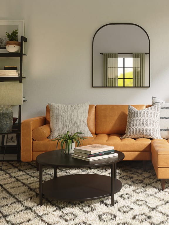 Brown leather couch with white and cream throw pillows. In front of the couch is a round coffee table with books and a plant resting on it. A white rug with a balck desgin is under the couch.