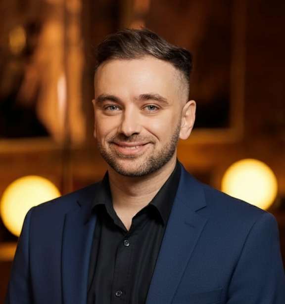 A man in a blue suit and black shirt stands in front of a blurred background.