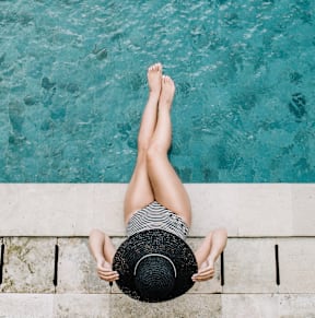 A woman in a striped shirt and black hat is sitting on the edge of a pool.