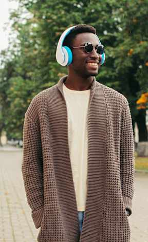 A man in a brown coat and sunglasses is wearing headphones and standing on a sidewalk.