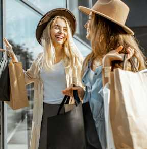 Two women wearing hats and carrying shopping bags are standing outside a store.