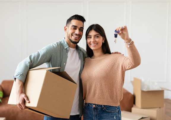 A man and woman are standing in a room with boxes, holding a key and a box.