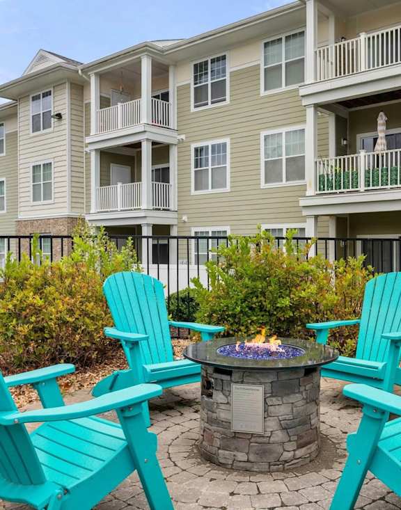 A fire pit surrounded by four blue Adirondack chairs in front of a building.