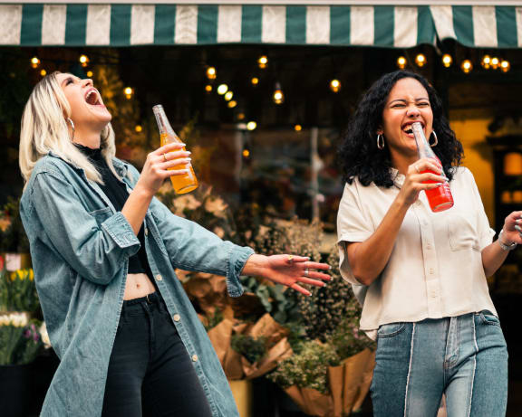 two women drinking beer and laughing while standing outside