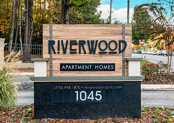A sign for Riverwood Apartment Homes is displayed in front of a wooded area.