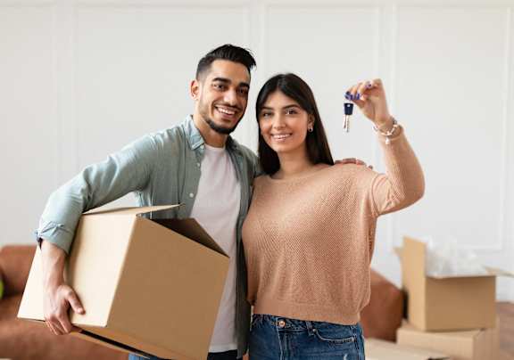 A man and woman are standing in a room with boxes, holding a key and a box.