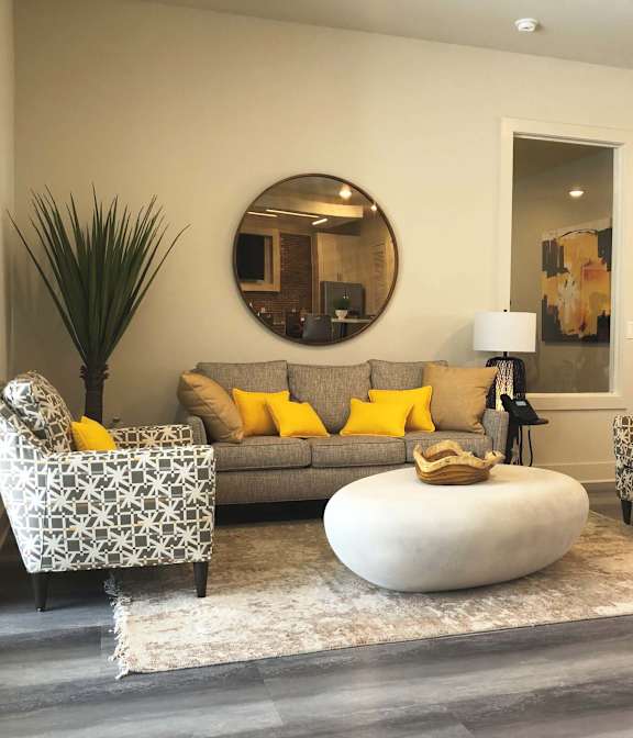 A living room with a grey couch, yellow pillows, a round mirror, and a white coffee table.