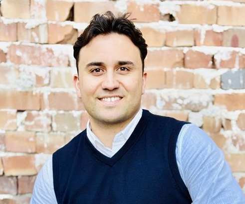 A man in a blue vest and light blue shirt is sitting in front of a brick wall.