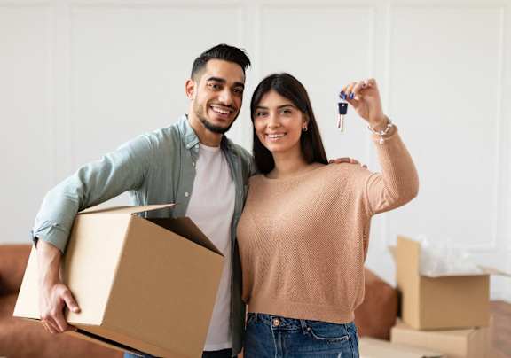 A man and woman are standing in a room with boxes, holding a key and a box.