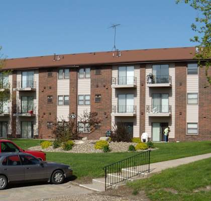 A grey car is parked in front of a red truck and a brown apartment building. at Southern Hills Apartments, Marshalltown, IA, 50158