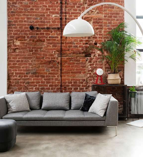 A grey sofa with a white lamp and a red vase on a table.