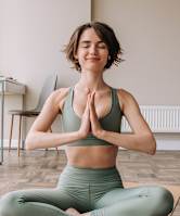 A woman in a green sports bra and leggings is meditating on a yoga mat in a bright room.