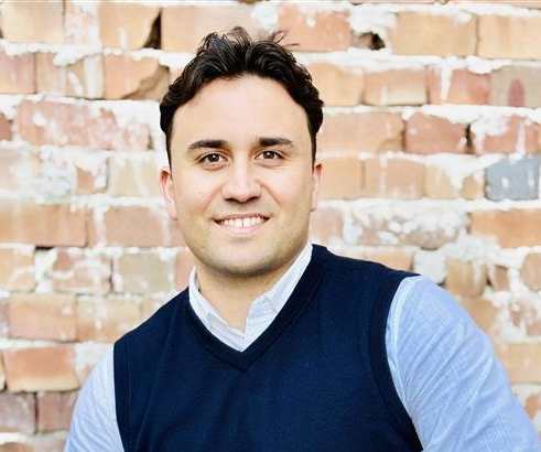 A man in a blue vest and light blue shirt is sitting in front of a brick wall.