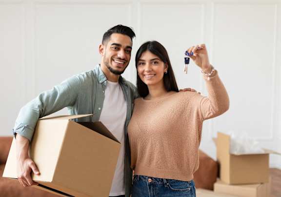 A man and woman are standing in a room with boxes, holding a key and a box.