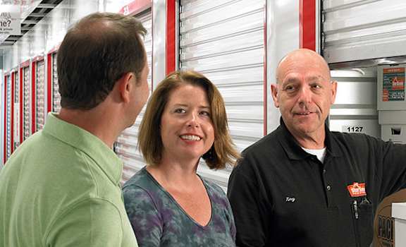 Three people in a storage unit with boxes and a sign that says "Heated and Temperature Controlled Storage units For what matters.".