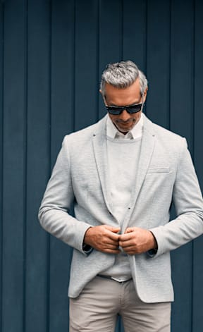 A man in a light grey suit and sunglasses is adjusting his jacket.