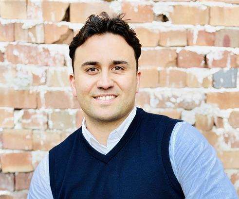 A man in a blue vest and light blue shirt is sitting in front of a brick wall.
