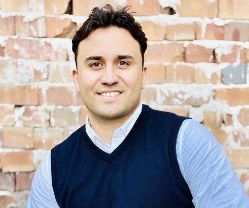 A man in a blue vest and light blue shirt is sitting in front of a brick wall.