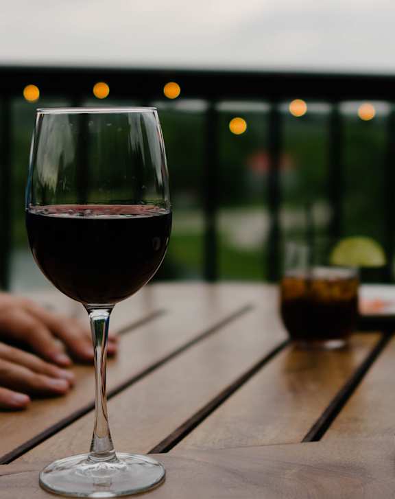 A glass of red wine is on a table with a person's hands in the background.