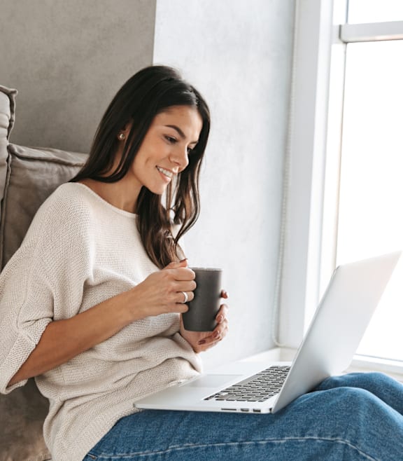 A woman is sitting on a couch with a laptop and a mug in her hands.