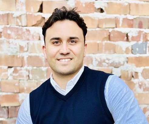 A man in a blue vest and light blue shirt is sitting in front of a brick wall.