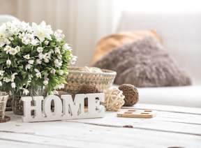 A white wooden table with a white flower pot, a white HOME sign, and a brown knitted pillow.