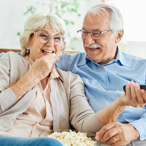 An elderly couple is sitting on a couch, laughing and holding a remote control.