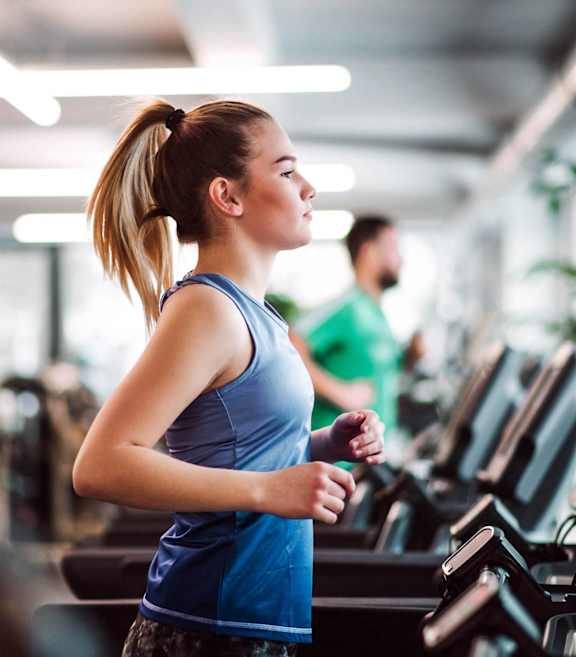 A woman in a blue top and grey shorts is running on a treadmill in a gym.