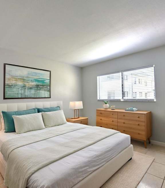 A bedroom with a large bed, a nightstand, a lamp, a dresser, and a potted plant.