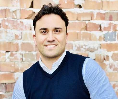 A man in a blue vest and light blue shirt is sitting in front of a brick wall.