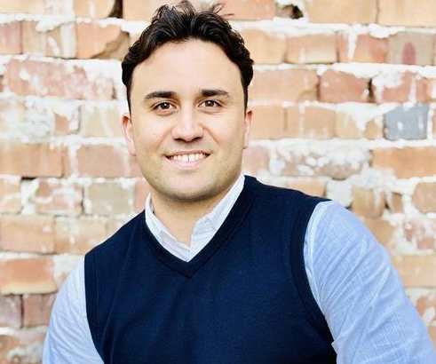 A man in a blue vest and light blue shirt is sitting in front of a brick wall.