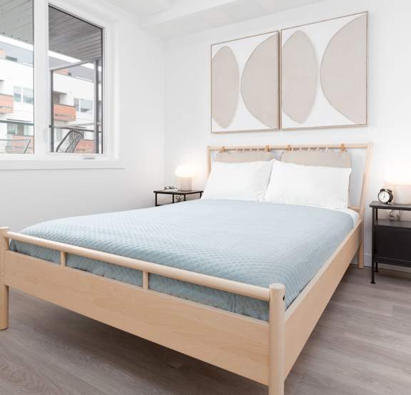 A bed with a blue sheet and two pillows sits in a room with a wooden frame and a nightstand with a lamp.