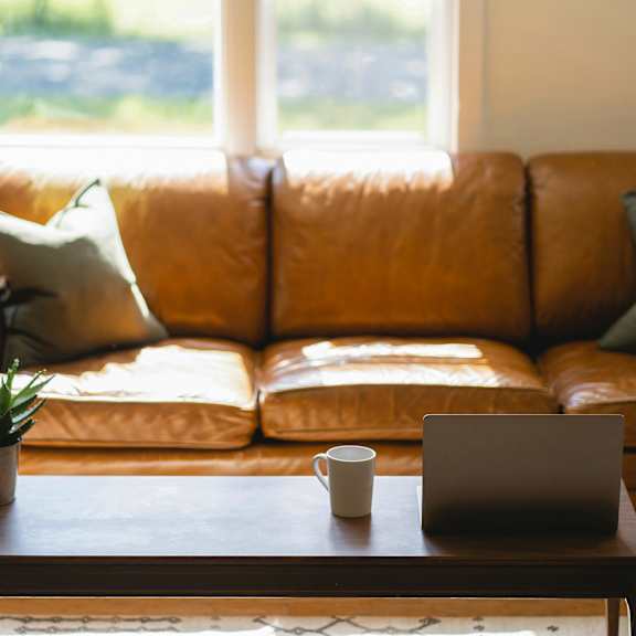 A brown leather couch with a laptop and a cup on a wooden table in front of it.