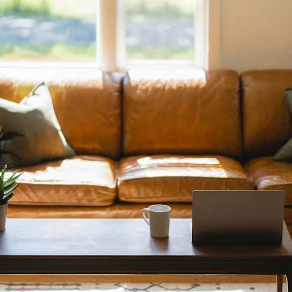 A brown leather couch with a laptop and a cup on a wooden table in front of it.