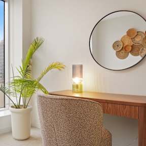 A room with a chair, a table, a plant, and a mirror with a decorative piece on it.