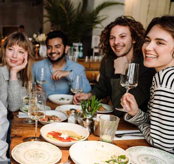 A group of people are sitting around a table with plates of food, taking a selfie with a smartphone.