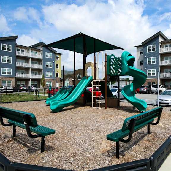 A playground with a green slide and two benches in front of apartment buildings.
