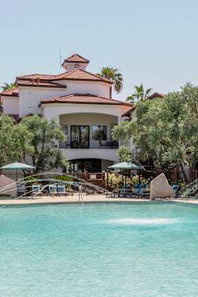 A large swimming pool in front of a house with a patio and palm trees.